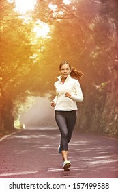 Young Woman Running. Female Runner Jogging On Misty Road With The Early Morning At Sunrise With Sun Breaking Through The Trees As She Trains During A Fitness Workout.