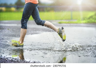 Young woman running on asphalt sports field in rainy weather. Details of legs and sports shoes splashing in puddles.