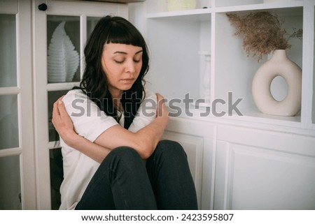 A young woman sadly sits in a corner and hugs herself in a bright room. The concept of the inner child and the protection of the inner world. A beautiful sad girl sits on the floor in secluded corner