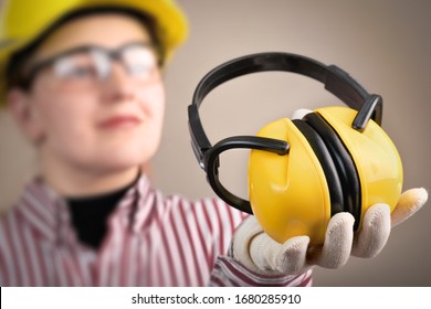 Junge Frau in Schutzbrille und Helm hält Kopfhörer in der Hand, um den Hörschutz zu gewährleisten: Konzept für Sicherheit und Gesundheitsschutz am Arbeitsplatz und Schutzausrüstung.