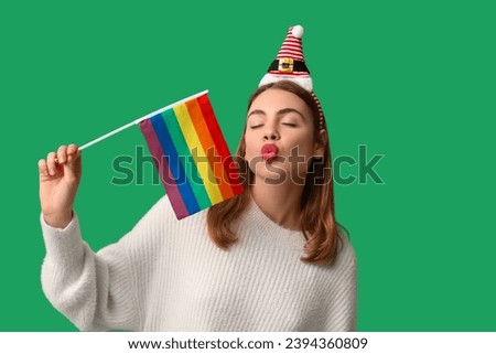 Young woman in Santa hat with LGBT flag blowing kiss on green background
