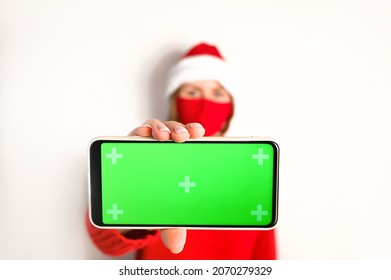 Young Woman In Santa Hat And Wearing Mask Holds Phone With Green Screen.Mock Up.Pandemic Christmas