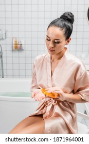 Young Woman In Satin Bathrobe Pouring Cosmetic Oil In Bathroom