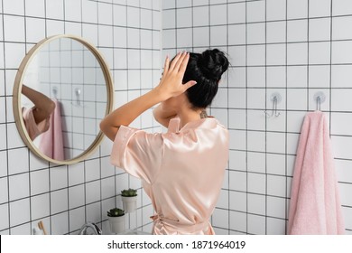 Young Woman In Satin Bathrobe Touching Hair Near Mirror In Bathroom