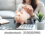 Young woman saving money monthly expenses putting coin in to piggy bank on the table.