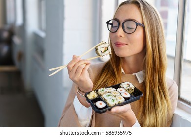 Young woman savoring her sushi lunch standing in front of a window in the office with her eyes closed and an expression of bliss as she anticipates the next mouthful