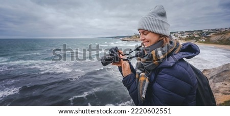 A young woman in a scarf jacket and a hat, a traveler professional photographer with a camera in her hands on the shore of the winter sea
