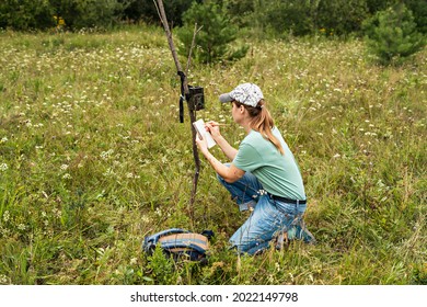 Young woman scientist zoologist writing down data from trap camera to notepad, observing wild animals in taiga forest, monitoring of rare and endangered animal species, World Wildlife Day ecology