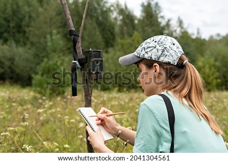 Young woman scientist zoologist writing down data from trap camera to notepad, observing wild animals in reserve taiga forest, monitoring of rare endangered animal species, ecology World Wildlife Day