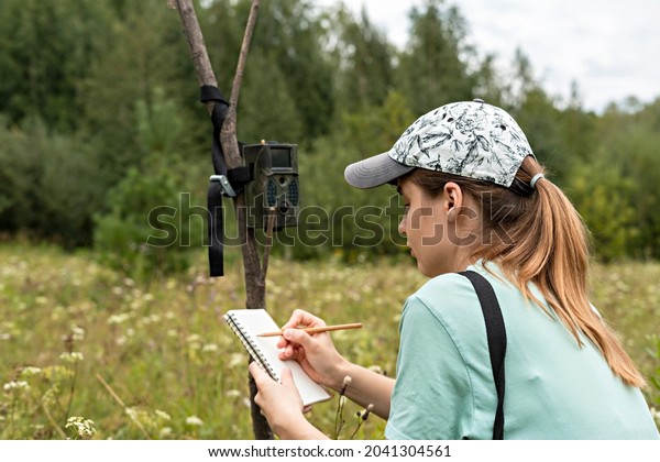 Young woman scientist zoologist writing down data\
from trap camera to notepad, observing wild animals in reserve\
taiga forest, monitoring of rare endangered animal species, ecology\
World Wildlife Day