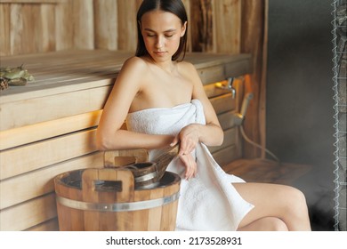 Young woman scoops up  water from a wooden bucket with a ladle in the sauna to create steam