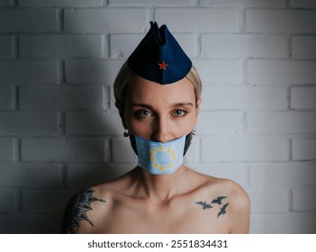 A young woman is seen in a powerful yet symbolic portrait, wearing a Yugoslavian pioneer hat, a European Union flag mask over her mouth, and showcasing bold tattoos on her shoulders. - Powered by Shutterstock