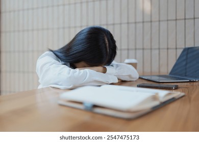 A young woman is seen resting her head on her arms at a wooden table in a cozy cafe, surrounded by a coffee cup and notebook, conveying a moment of fatigue and contemplation. - Powered by Shutterstock