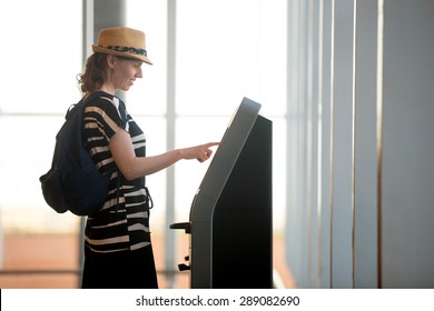 Young woman at self service transfer area doing self-check-in or buying plane tickets at automated machine with touchscreen interactive display in modern airport terminal building