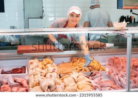 Young woman seller in uniform lays out chicken meat in showcase of butcher shop