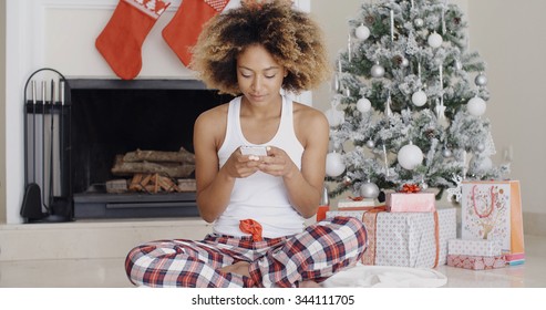 Young Woman Sending A Christmas Text Message On Her Mobile Phone As She Sits On The Floor In Front Of The Decorated Hearth And Xmas Tree.