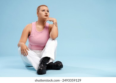 Young woman with a shaved head is sitting on the floor with her hand on her chin, looking thoughtfully into the distance - Powered by Shutterstock