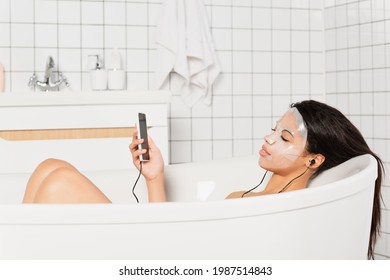 young woman with sheet mask on face lying with earphones and cellphone in bathtub - Powered by Shutterstock