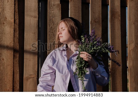 Young woman in shirt with closed eyes holding bouquet of purple flowers standing alone in front of wooden fence enjoying of sunlight at golden hour. Summer outfit. Girl with flowers