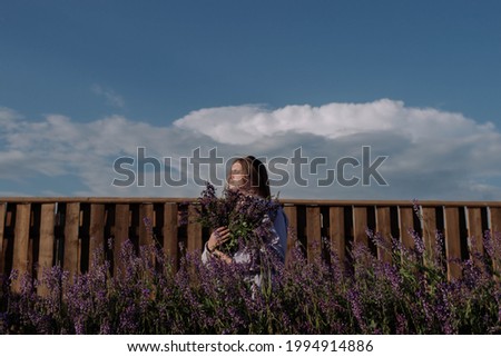 Young woman in shirt holding bouquet of purple flowers standing alone among field in front wooden fence and blue sky enjoying of sunlight at golden hour. Summer outfit. Girl with flowers