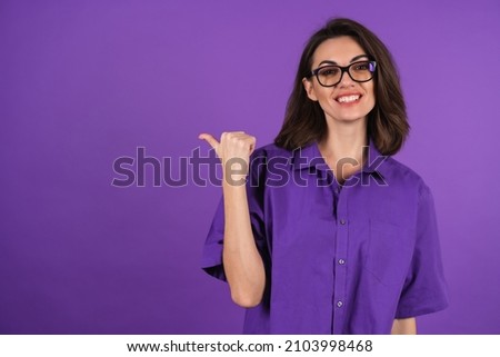 Young woman in a shirt with short sleeves on a purple background, with makeup and stylish glasses, points a finger to the left with a smile