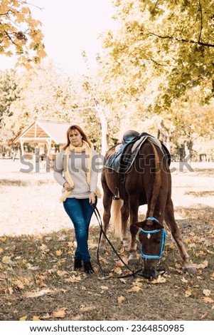 A young woman in a shirt stands next to a brown horse. Horseback riding in the autumn forest. High quality photo