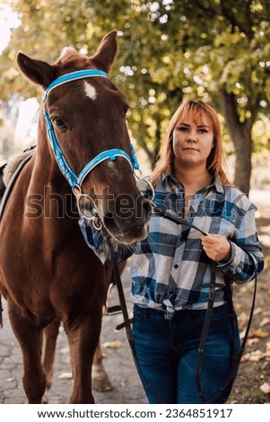 A young woman in a shirt stands next to a brown horse. Horseback riding in the autumn forest. High quality photo