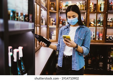 Young Woman Shopper Wearing Face Mask To Protect From Virus Holding Bottle Of Wine And Looking Into Screen Of Her Smart Phone While Standing Near Shelf In Supermarket.