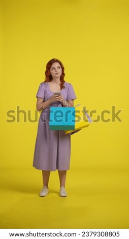 Young woman with shopping bags uses smartphone. A woman looks through lucrative offers, promotions, and makes an online order. Full length redhaired woman in studio on yellow background. Vertical shot