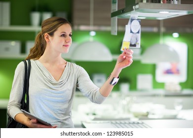 Young Woman Shopping For Furniture In A Furniture Store, Using Her Tablet Computer To Compare Prices/check For Dimensions