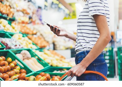 Young woman shopping healthy food in supermarket blur background. Female hands buy nature products using smart phone in store. Hipster at grocery using mobile. Person comparing price of produce - Shutterstock ID 609060587