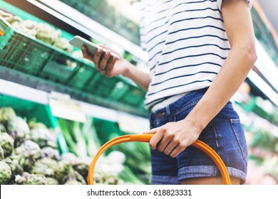 Young Woman Shopping Healthy Food In Supermarket Blur Background. Female Hands Buy Products And Using Mobile Smart Phone In Store. Hipster At Grocery Holding Basket. Person Comparing Price Of Produce