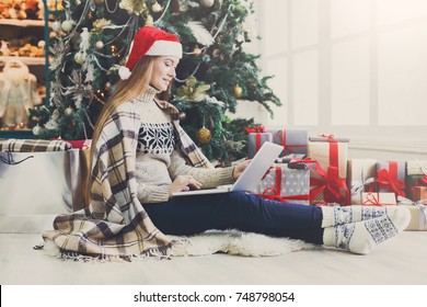 Young woman shopping online on laptop in cozy christmas interior. Girl in casual sitting under decorated pine tree among lots of wrapped presents. Preparing to xmas, bying on winter sales. Copy space ภาพถ่ายสต็อก