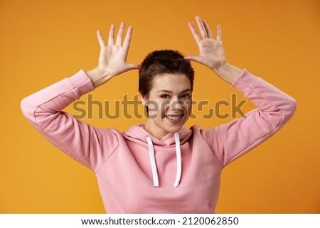 Young woman with short hair doing funny gesture over head as bull horns over yellow background