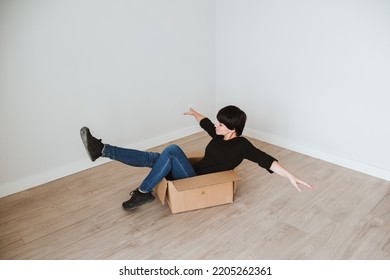 A Young Woman With Short Hair Inside A Cardboard Box In An Empty Room. Concept Of Moving And Furnishing A New Home. Solitary Life. Become Independent. Single Woman