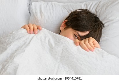 Young Woman With Short Hair Peeks Out From Under The Covers Waking Up In Bed In The Morning. Hide And Seek Behind The Blanket. Healthy Sleep.