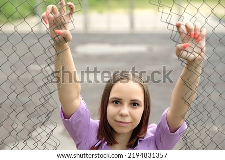 Young woman with short hair in purple shirt posing, holding on to bars with hands, looking through hole of lattice fence