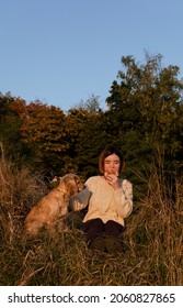 Young Woman With Short Hair Sitting On Dry Grass Hill During Sunset With Her English Cocker Spaniel Dog, Eating Pastry.