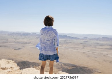 A young woman with short hair stands on a rocky cliff, wearing a light shirt and denim dress, looking out over an expansive desert with distant mountains and clear skies. - Powered by Shutterstock