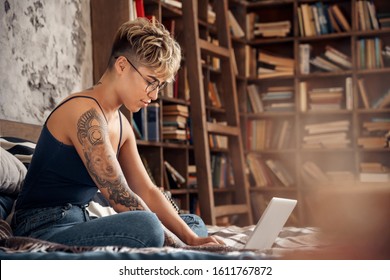 Young woman short hair wearing eyeglasses sitting on bed with laptop working online side hustle smiling joyful