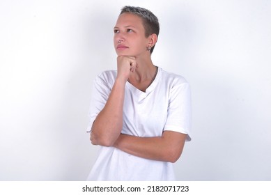 young woman with short hair wearing white t-shirt over white background with hand under chin and looking sideways with doubtful and skeptical expression, suspect and doubt. - Powered by Shutterstock