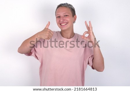 young woman with short hair wearing pink t-shirt over white background  smiling and looking happy, carefree and positive, gesturing victory or peace with one hand
