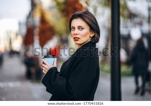Young woman with short haircut and red lips, holds\
a cup of coffee, strolling through the streets of the city. Around\
are people and cars. 