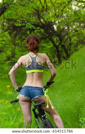 Young woman in shorts riding a bicycle, view from the back. A sports woman in short shorts and tops sits on a bicycle, a bright summer photo in the forest.