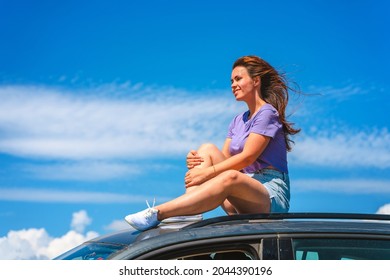 A young woman in shorts is sitting on the roof of a Volkswagen Tiguan car with a view of a beautiful blue sky with clouds. The concept of traveling by car.