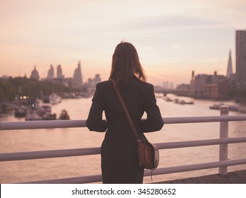 A Young Woman With A Shoulder Bag Is Standing On A Bridge And Is Admiring The Sunrise Over The London Skyline