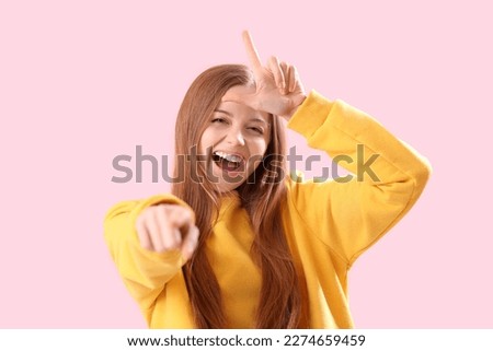 Young woman showing loser gesture and pointing at viewer on pink background, closeup