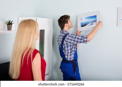 Young Woman Showing Male Carpenter For Hanging Picture Frame On Wall At Home