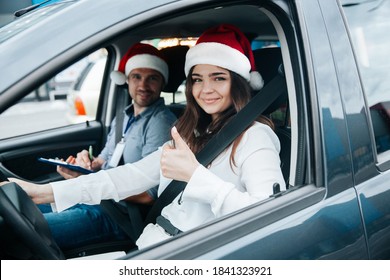 Young Woman Showing Thumb Up. Merry Christmas And Happy New Year Driving Courses On Winter Holidays. Female Student And Instructor In Santa Hats Smiling And Looking Into Camera Through Car Window.