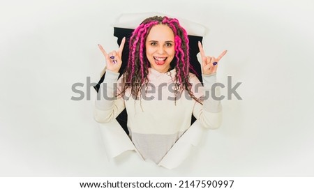 Young woman showing tongue and gesture of goat. Playful female with curly hairstyle sticking out of hole of white background